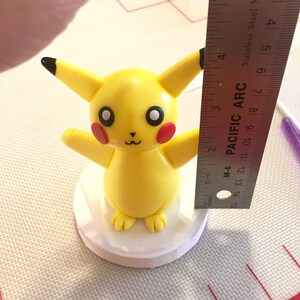 May include: A yellow Pikachu figurine with black-tipped ears, red cheeks, and a smiling face. The figurine is on a white base, and a ruler shows it is approximately 7.6 cm tall. The figurine is on a white base.