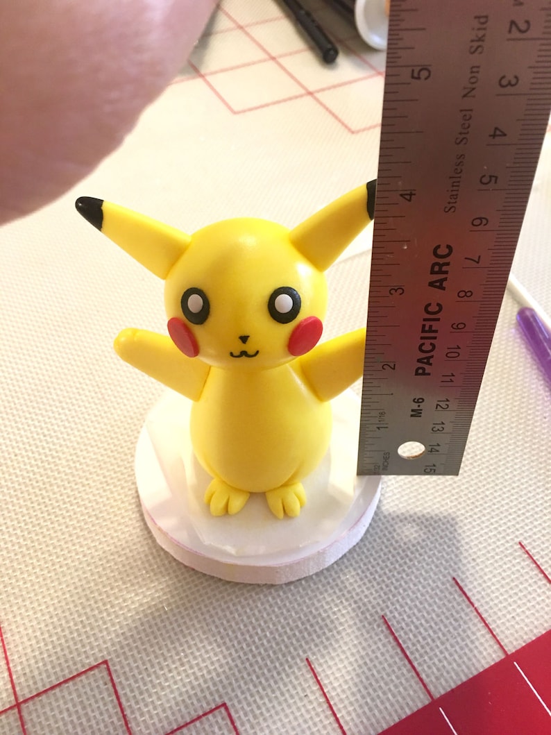 May include: A yellow Pikachu figurine with black-tipped ears, red cheeks, and a smiling face. The figurine is on a white base, and a ruler shows it is approximately 7.6 cm tall. The figurine is on a white base.