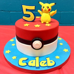 May include: A colourful birthday cake featuring a Pikachu figure and the number 5. The cake is decorated in red, white, and blue, with yellow stars and the name "Caleb" on the base. The cake sits on a red surface.