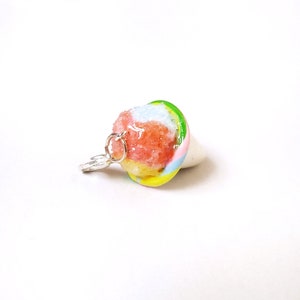 Puede incluir: Un amuleto de arcilla polimérica hecho a mano de colores en forma de cono de hielo raspado con un anillo de salto plateado. El hielo raspado es rosa, azul y amarillo con un borde verde.