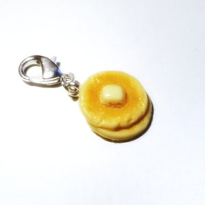 Könnte beinhalten: Ein silberner Anhänger mit einem Miniaturstapel von zwei Pfannkuchen mit einem Stück Butter darauf.