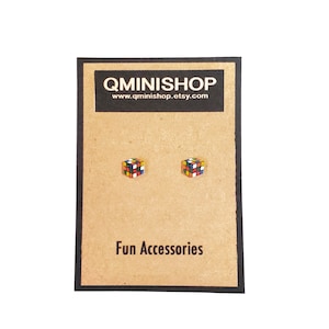 Może przedstawiać: Para złotych kolczyków w kształcie miniaturowej kostki Rubika z kolorowymi kwadratami. Kolczyki są prezentowane na brązowej kartce z tekstem "QMINISHOP" i "www.qminishop.etsy.com". Na kartce widnieje również napis "Fun Accessories".