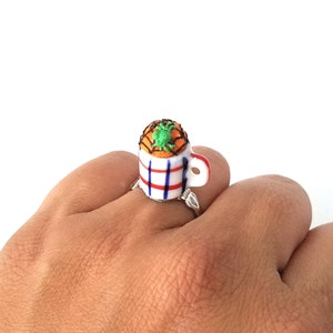 Könnte beinhalten: Ein weißer Keramikring mit blau-rotem Streifenmuster. Der Ring zeigt eine Miniaturtasse mit einer kleinen orangefarbenen und grünen Spinne darauf.