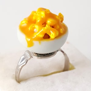 May include: A white ceramic ring with a yellow mac and cheese design. The ring is made to look like a small bowl of mac and cheese.