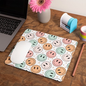 May include: A white mouse pad with a pattern of colorful smiley faces and gold stars. The mouse pad is on a wooden desk with a laptop, a pencil, a vase of pink flowers, and a tape dispenser.
