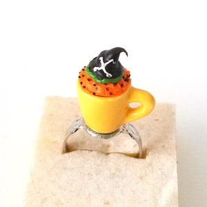 Könnte beinhalten: Ein gelber Keramik-Teetassenring mit einem Halloween-Cupcake darauf. Der Cupcake ist mit einem schwarzen Hexenhut mit einem weißen Totenkopf und Knochen-Design verziert. Der Cupcake ist mit orangefarbenem, grünem und schwarzem Frosting dekoriert.