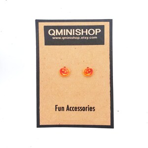 Può includere: Un paio di orecchini a bottone a forma di zucca di Halloween arancioni e rossi su una carta marrone con il testo "QMINISHOP" e "www.qminishop.etsy.com". La carta dice anche "Fun Accessories".