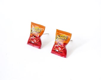 Può includere: Un paio di orecchini con bustine di Cheetos in miniatura. Le bustine sono rosse e arancioni con il logo Cheetos e le parole "Crunchy" stampate su di esse.