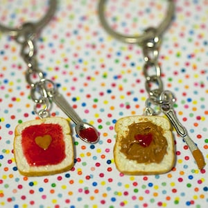 Op de afbeelding: Twee sleutelhangers met miniatuur pindakaas- en jam-sandwiches. Elk sandwich heeft een hartvormig stukje jam of pindakaas in het midden. De sleutelhangers zijn bevestigd aan zilveren lepels.