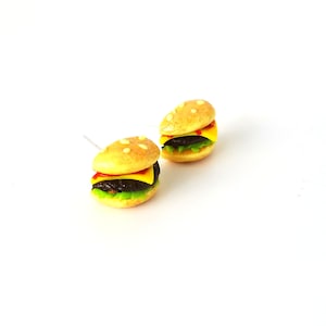 May include: A pair of polymer clay earrings shaped like miniature cheeseburgers. Each earring features a brown patty, yellow cheese, red ketchup, green lettuce, and a sesame seed bun.