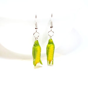 May include: A pair of silver dangle earrings featuring a realistic corn on the cob design. The corn is green with yellow kernels and has a silver hook.