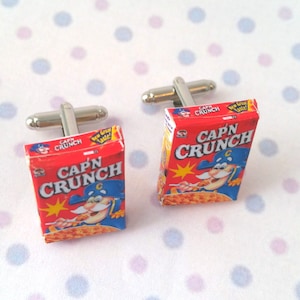 May include: A pair of cufflinks featuring miniature cereal boxes with the Cap'n Crunch logo and mascot. The boxes are red and white with a yellow star and the text "Cap'n Crunch" in bold, white letters.