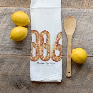 May include: A white kitchen towel with a watercolor illustration of three soft pretzels. The text "Philadelphia Soft Pretzel" is printed below the pretzels. The towel is lying on a wooden surface with lemons and a wooden spoon.