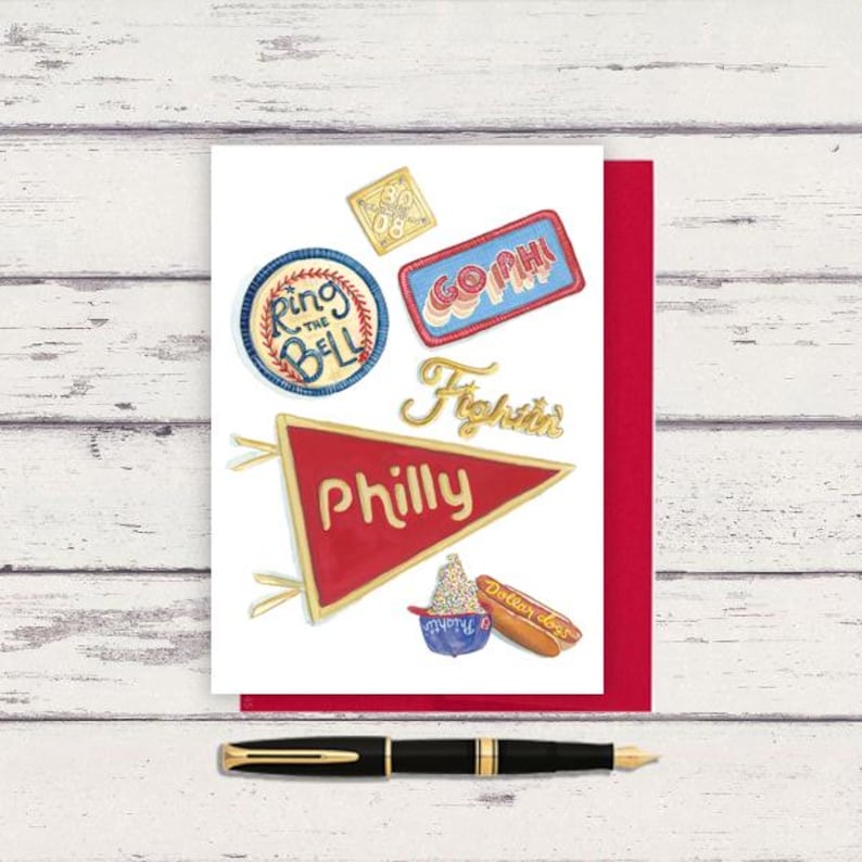 May include: A greeting card with a red border featuring watercolor illustrations of Philadelphia-themed icons. The card includes a pennant that says "Philly", a hot dog, and other patches with text. A black pen with gold accents rests below.