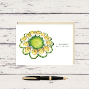 May include: Greeting card with a watercolor illustration of deviled eggs arranged in a circle. The card has the text "Let's Celebrate, You Tasty Devil". A black pen with gold accents is in the foreground.
