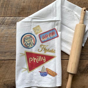 May include: White kitchen towel with embroidered patches and a wooden rolling pin. The towel features patches with the words "Ring the Bell", "Go Phils", and "Philly". A red pennant with "Philly" is also visible. The rolling pin is made of light-colored wood.