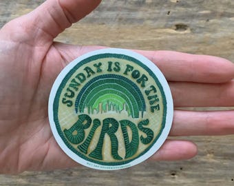 May include: A round sticker with the text "SUNDAY IS FOR THE BIRDS" in green, arched around the top and bottom. The center features a green rainbow over a city skyline. The sticker is held in a hand.