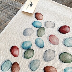 Op de afbeelding: Een wit canvas placemat met een patroon van kleurrijke eieren. De eieren zijn geschilderd in tinten blauw, groen, bruin en roze. De placemat heeft een label eraan bevestigd met de tekst "Meadowbrook Lane".