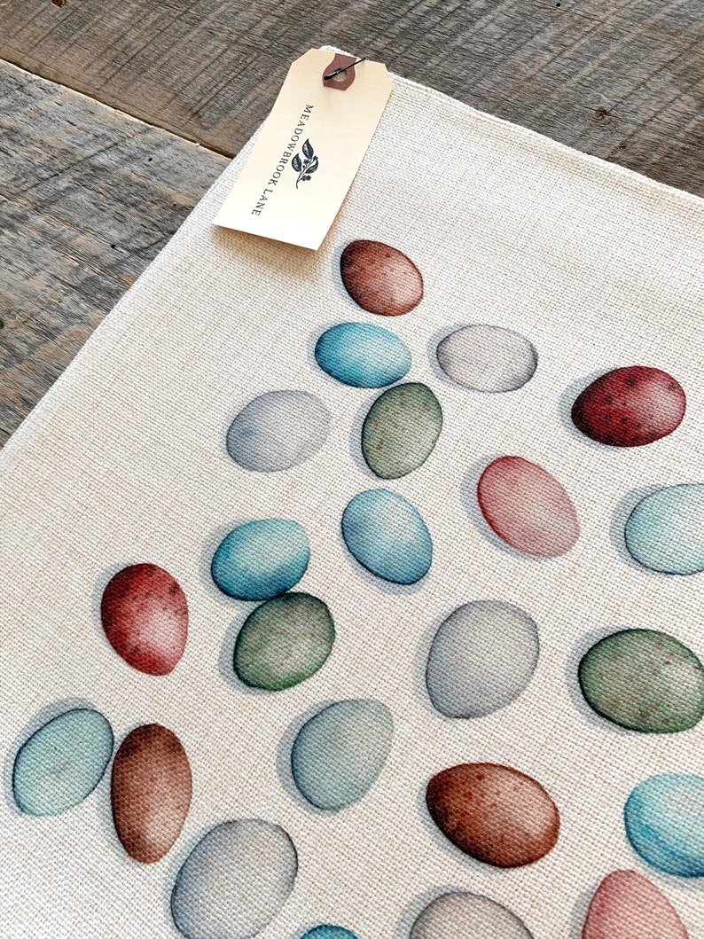 Op de afbeelding: Een wit canvas placemat met een patroon van kleurrijke eieren. De eieren zijn geschilderd in tinten blauw, groen, bruin en roze. De placemat heeft een label eraan bevestigd met de tekst "Meadowbrook Lane".