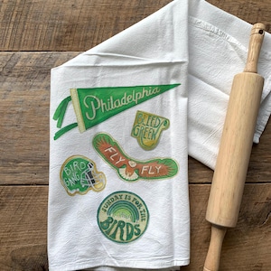 May include: White kitchen towel with green and gold embroidered patches. The patches feature the words "Philadelphia", "Fly", "Bird Gang", "Bleed Green", and "Sunday is for the Birds".