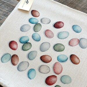 Op de afbeelding: Een witte linnen placemat met een patroon van kleurrijke eieren. De eieren zijn geschilderd in tinten blauw, groen, bruin en roze. De placemat heeft een label met de tekst "Meadowbrook Lane".