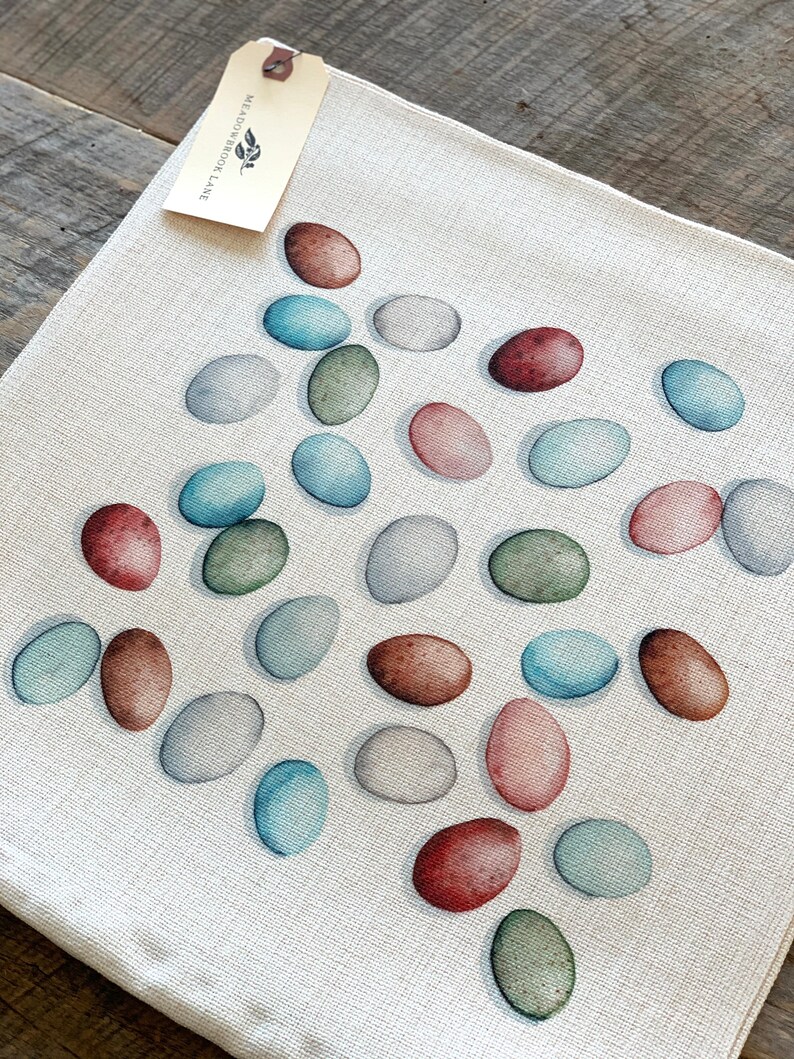 Op de afbeelding: Een witte linnen placemat met een patroon van kleurrijke eieren. De eieren zijn geschilderd in tinten blauw, groen, bruin en roze. De placemat heeft een label met de tekst "Meadowbrook Lane".