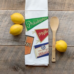 May include: White kitchen towel with pennant designs that read "Philadelphia" and "Philly". The towel also features a basketball design and the text "Here They Come Philadelphia". Three lemons and a wooden spoon are next to the towel.