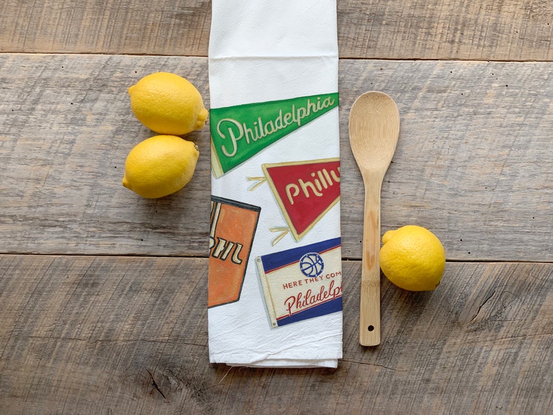May include: White kitchen towel with pennant designs that read "Philadelphia" and "Philly". The towel also features a basketball design and the text "Here They Come Philadelphia". Three lemons and a wooden spoon are next to the towel.