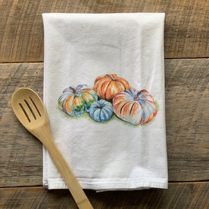May include: A white kitchen towel with a colorful watercolor illustration of four pumpkins. The pumpkins are orange, green, blue, and white.