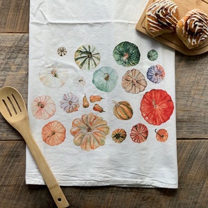 May include: White kitchen towel featuring watercolour illustrations of pumpkins in orange, green, and blue. A wooden spoon is on the left, and two cinnamon rolls sit on a wooden board.