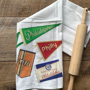 May include: White kitchen towel with vintage-style Philadelphia pennant designs in green, red, orange, and blue. The towel features the words "Philadelphia", "Philly", and "Here They Come Philadelphia". A wooden rolling pin rests on the towel.