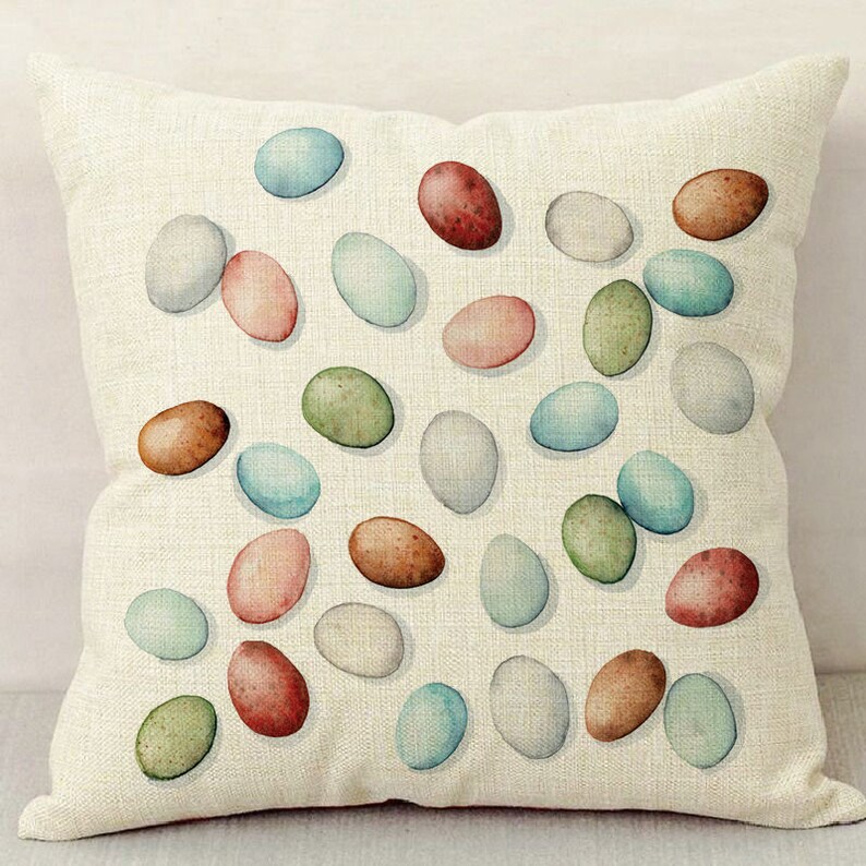 Op de afbeelding: Een witte linnen kussenhoes met een patroon van kleurrijke aquarel eieren. De eieren zijn in verschillende tinten blauw, groen, roze en bruin.