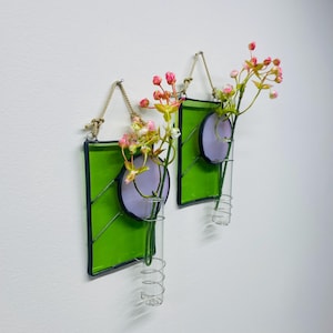 May include: Two green stained glass wall vases with a purple circle design and a clear glass tube for flowers. The vases are hung on a white wall with pink and white flowers in the tubes.