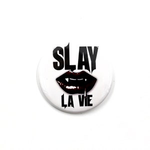 Könnte beinhalten: Ein weißer Knopf mit schwarzem Text, der "SLAY LA VIE" sagt, und einem schwarzen und roten Vampirmund mit Zähnen und tropfendem Blut.