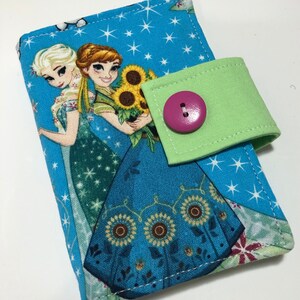 Puede incluir: Cartera de tela con fondo azul y estrellas blancas, con imágenes de dos personajes animados. Tiene una solapa verde con un botón rosa. Los personajes visten trajes azules y verdes.