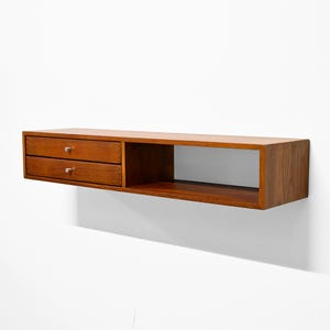 May include: A floating wooden shelf with a warm brown finish. It features two drawers on the left side with small, round knobs and an open compartment on the right. The shelf is mounted on a white wall.