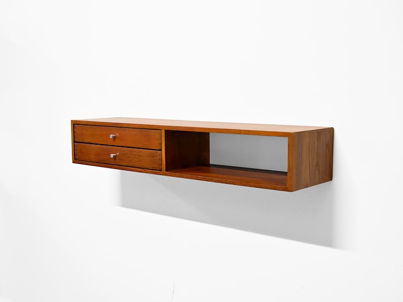 May include: A floating wooden shelf with a warm brown finish. It features two drawers on the left side with small, round knobs and an open compartment on the right. The shelf is mounted on a white wall.