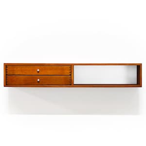 May include: A wall-mounted wooden shelf with two drawers and a rectangular open space. The drawers are a warm brown color with small, round silver knobs. The open space is white, and the shelf is mounted on a white wall.