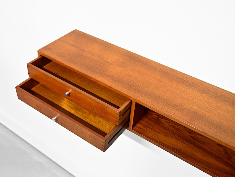 May include: A long, rectangular wooden shelf with two drawers and a lower open shelf. The drawers are open, revealing the interior. The wood has a warm, reddish-brown tone, and the hardware is silver. The shelf is set against a white background.