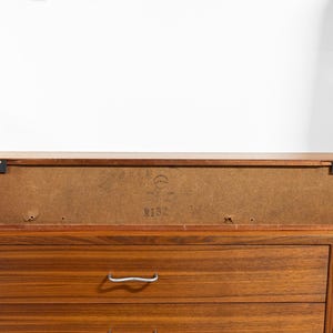 May include: A mid-century modern wooden cabinet with a rectangular shape. The cabinet is a warm brown color and features a drawer with a silver handle. The back of the cabinet has a label with the number "N132".