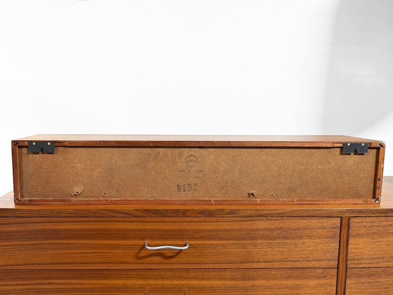 May include: A mid-century modern wooden cabinet with a rectangular shape. The cabinet is a warm brown color and features a drawer with a silver handle. The back of the cabinet has a label with the number "N132".