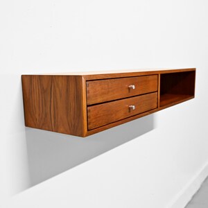 May include: A floating wooden shelf with two drawers and a cubby. The shelf is made of a warm-toned wood and has silver drawer pulls. The design is minimalist and modern, suitable for storage or display.