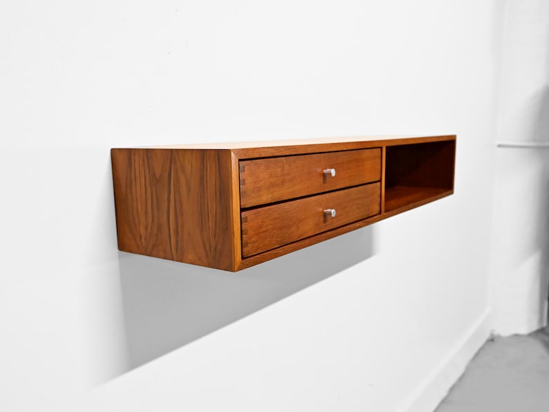 May include: A floating wooden shelf with two drawers and a cubby. The shelf is made of a warm-toned wood and has silver drawer pulls. The design is minimalist and modern, suitable for storage or display.