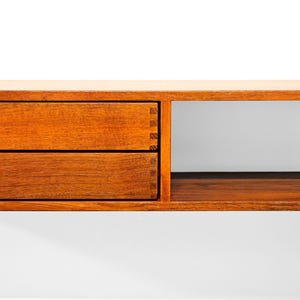 May include: A wooden floating shelf with two drawers and a cubby. The drawers are a warm brown with silver knobs. The shelf is made of wood and has a simple, modern design. The shelf is mounted on a white wall.