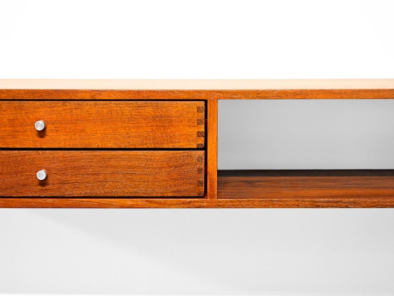 May include: A wooden floating shelf with two drawers and a cubby. The drawers are a warm brown with silver knobs. The shelf is made of wood and has a simple, modern design. The shelf is mounted on a white wall.