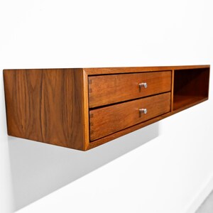 May include: A floating wooden cabinet with two drawers and a small open shelf. The cabinet is made of a warm-toned wood, with silver drawer pulls. The design is modern and minimalist, suitable for storage.