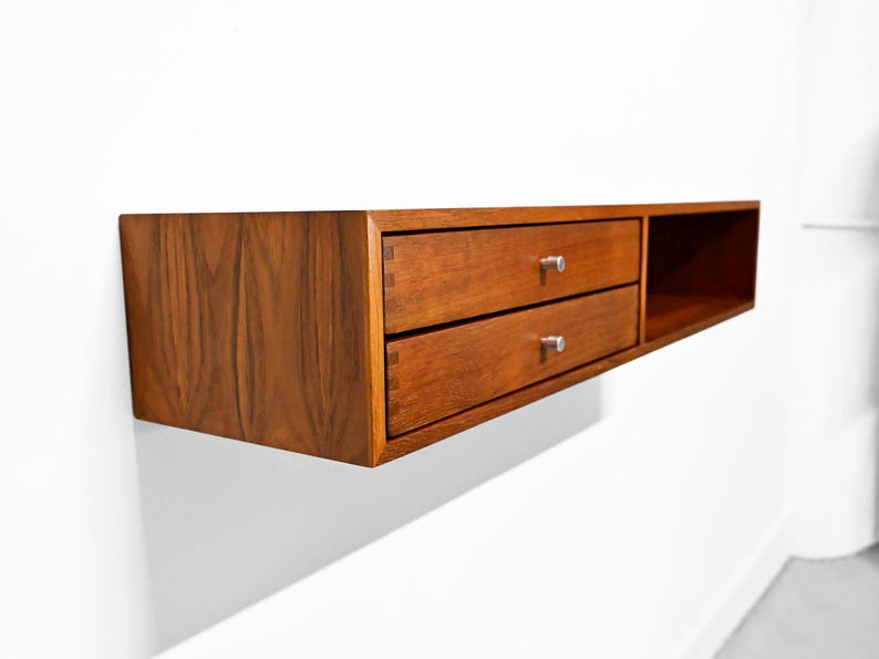 May include: A floating wooden cabinet with two drawers and a small open shelf. The cabinet is made of a warm-toned wood, with silver drawer pulls. The design is modern and minimalist, suitable for storage.