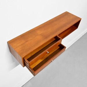 May include: A mid-century modern floating wall-mounted cabinet made of wood. The cabinet has a rectangular shape with two drawers and a shelf. The wood has a warm, reddish-brown tone. The drawers have small, silver-colored knobs.
