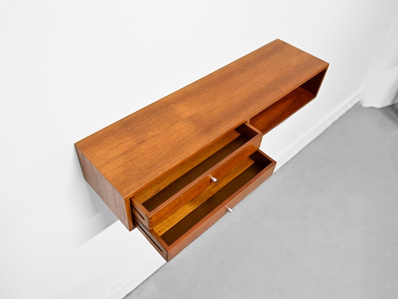 May include: A mid-century modern floating wall-mounted cabinet made of wood. The cabinet has a rectangular shape with two drawers and a shelf. The wood has a warm, reddish-brown tone. The drawers have small, silver-colored knobs.