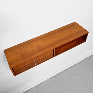 May include: A rectangular, wall-mounted wooden shelf with a warm brown finish. The shelf features two drawers with silver handles on the left and an open compartment on the right. The shelf is mounted on a white wall.